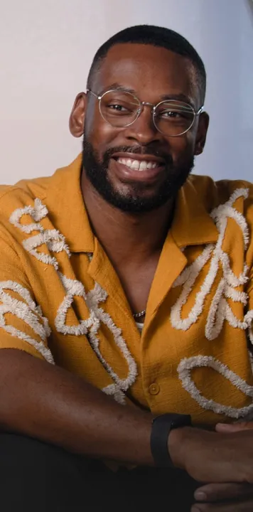 Smiling man wearing round glasses and a mustard yellow shirt with white textured patterns.